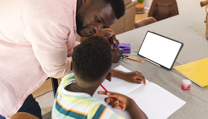 African American Father Assists A Young Son With Homework At A Table With Copy Space. Father And Son Engage In Educational Activities, With A Tablet And Stationery Items Present.